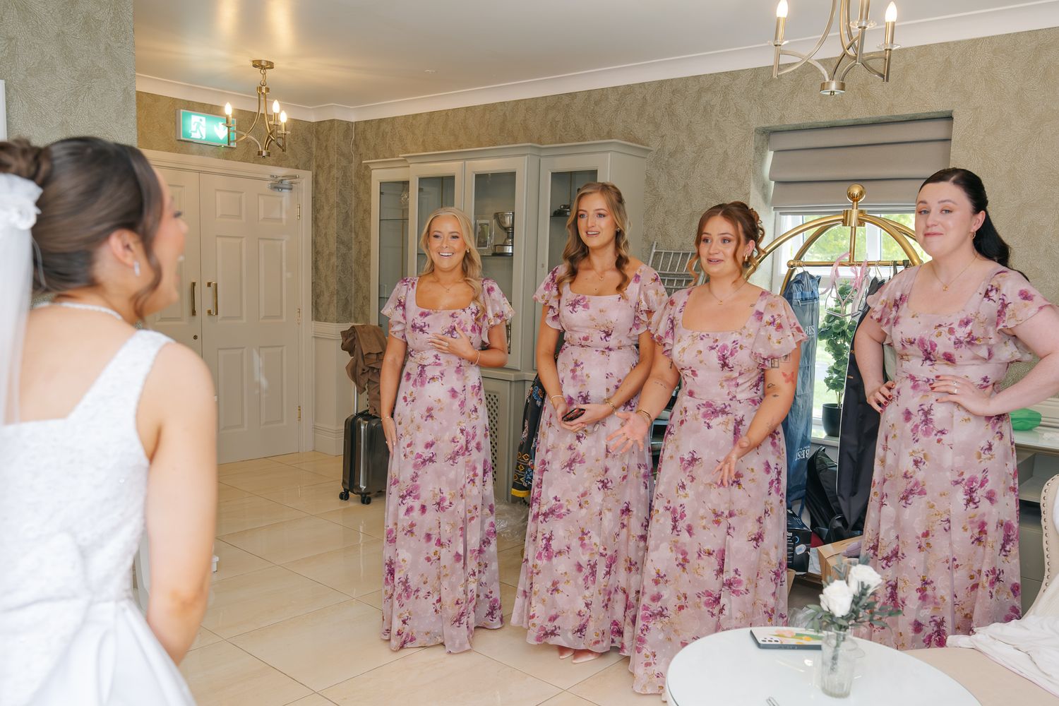 Bride reveals wedding dress to bridesmaids in floral pink gowns in elegant room.