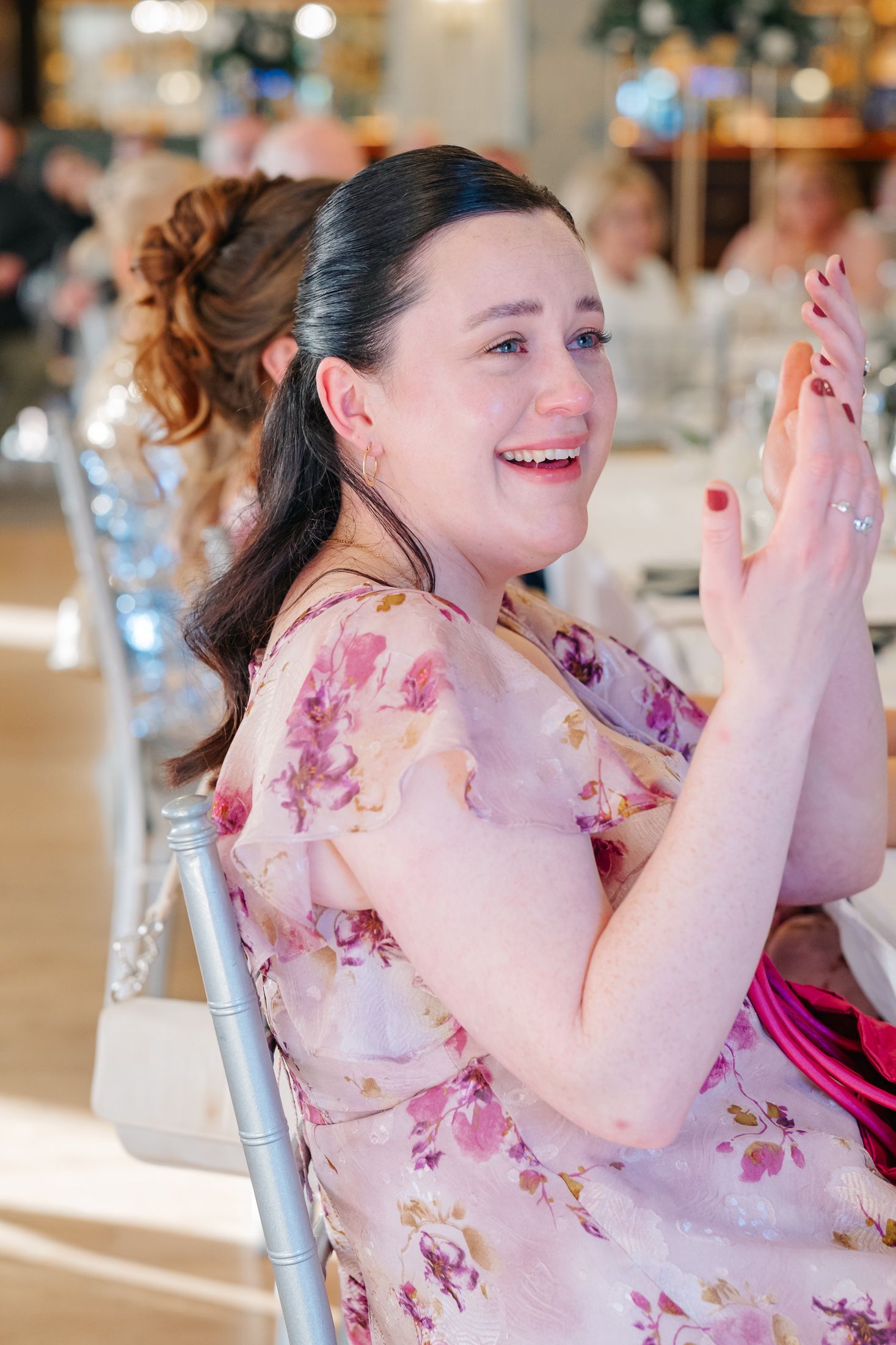 A woman in a floral dress laughs and claps at a wedding reception, seated at an elegantly decorated table.