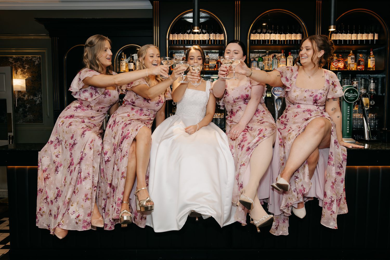 Bride and four bridesmaids in floral dresses toasting champagne while seated at a bar, laughing and kicking up heels.