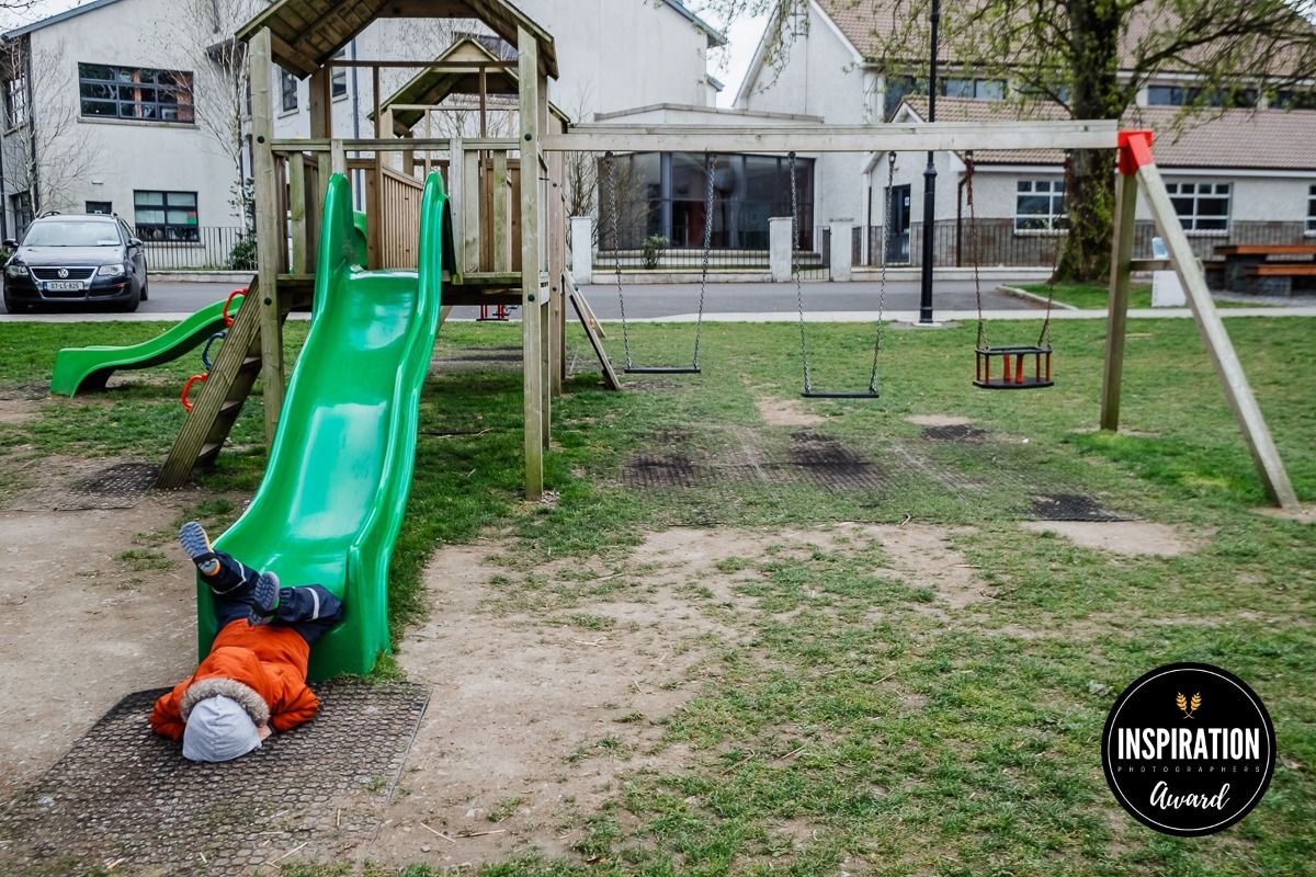 A bright green playground slide stands in a grassy park area with residential buildings visible in the background.