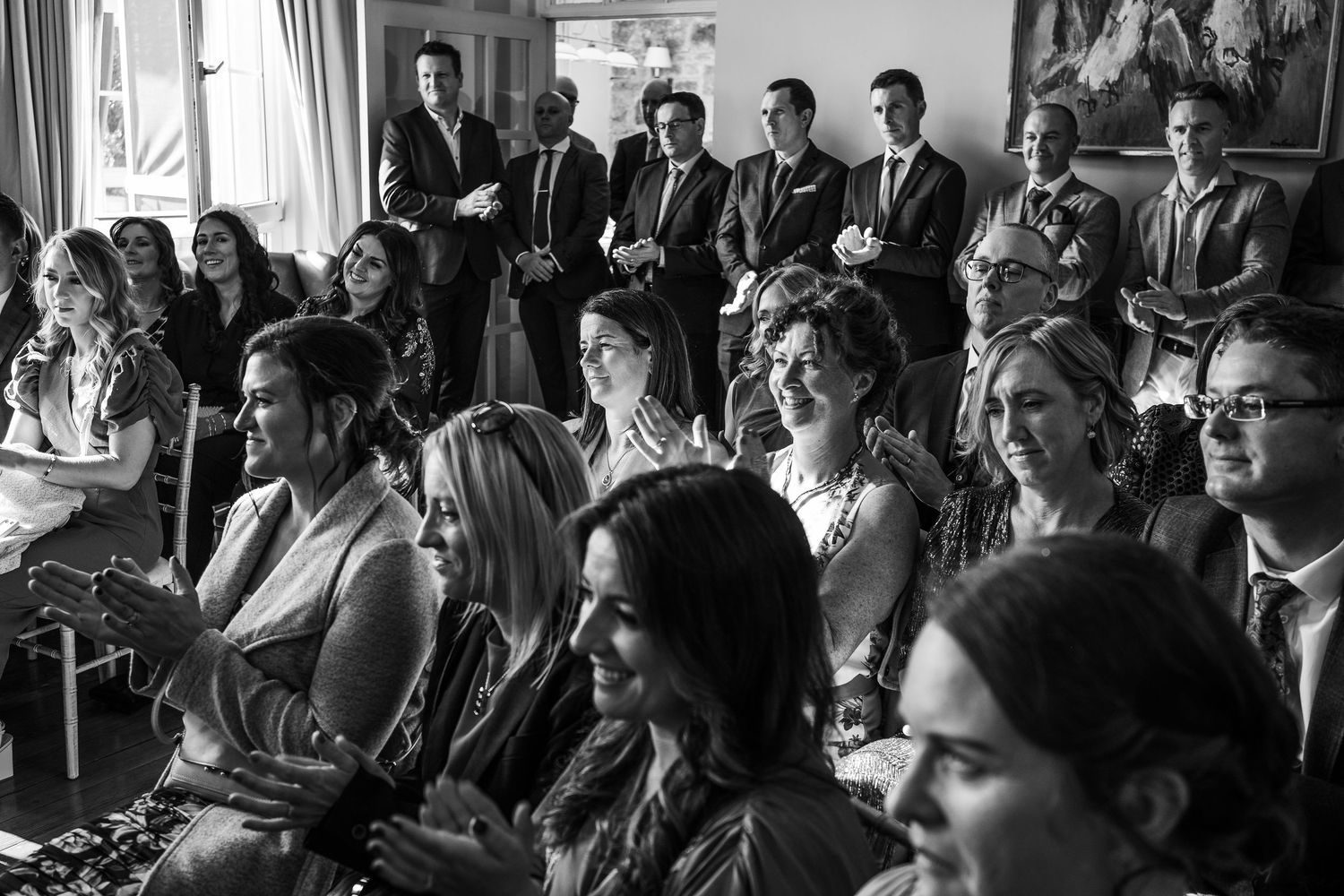 Black and white photo of wedding guests smiling and clapping during an indoor ceremony.