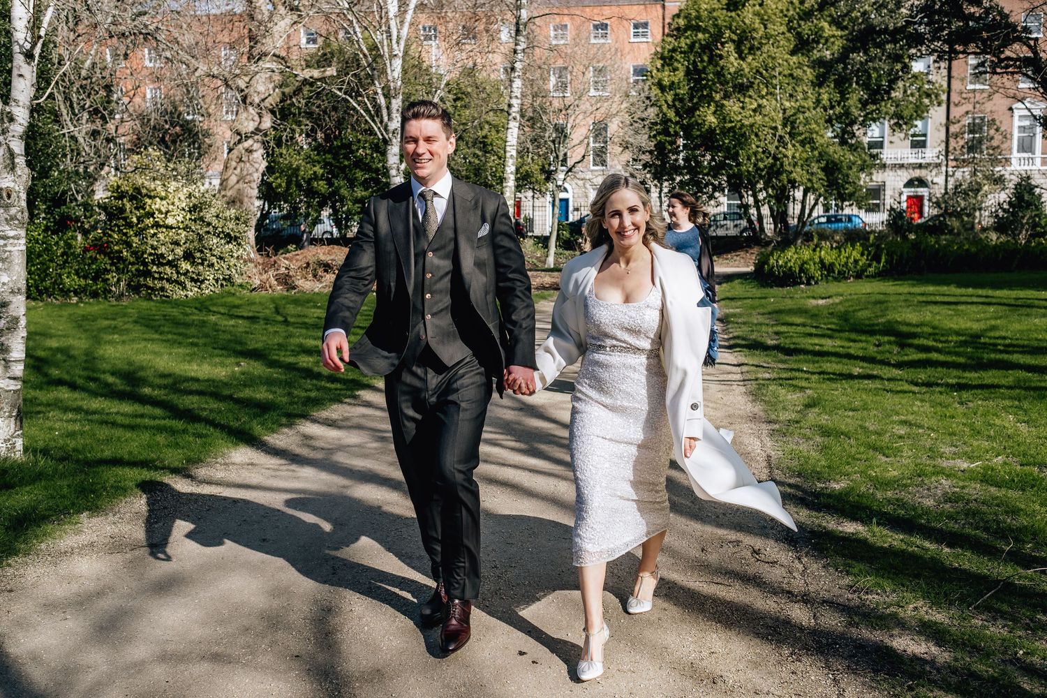 A joyful bride and groom walk hand in hand through a sunny park on their wedding day.