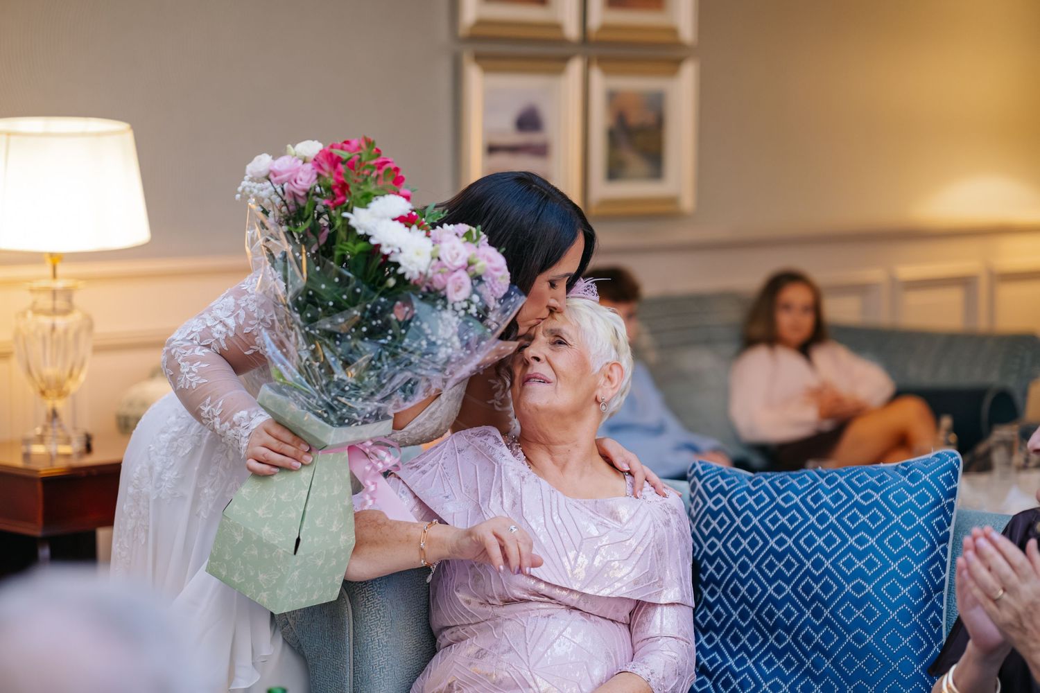 Young woman kisses elderly woman while presenting pink flower bouquet in cozy living room setting.