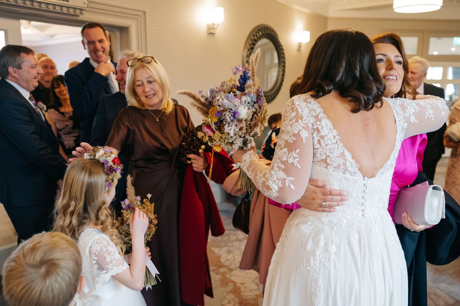 Bride in lace backless gown receives guests at wedding reception, surrounded by smiling family and flower girls.
