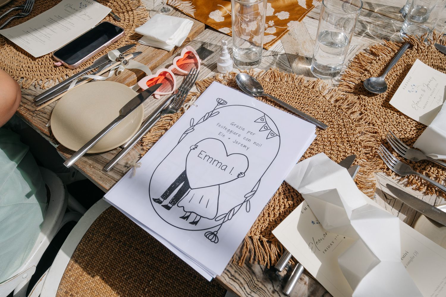 Wedding table setting with woven placemats, silverware, glasses, and a personalized illustrated place card reading Emma.