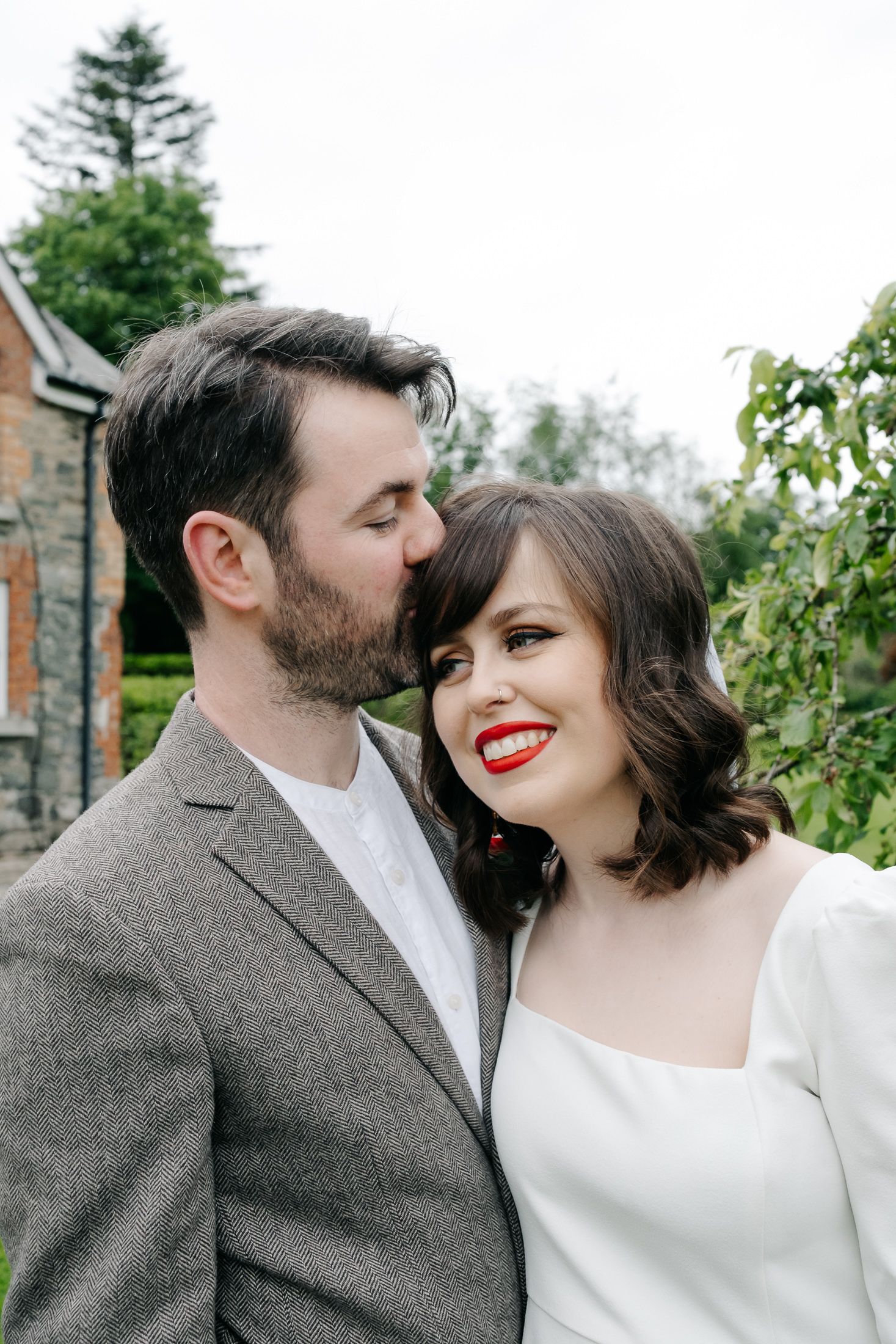 A groom in a grey suit kisses his bride on the cheek as she smiles wearing red lipstick and a white dress.