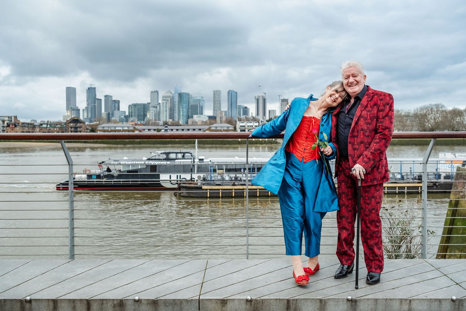 Two people in colorful suits pose together on waterfront with urban skyline behind them.