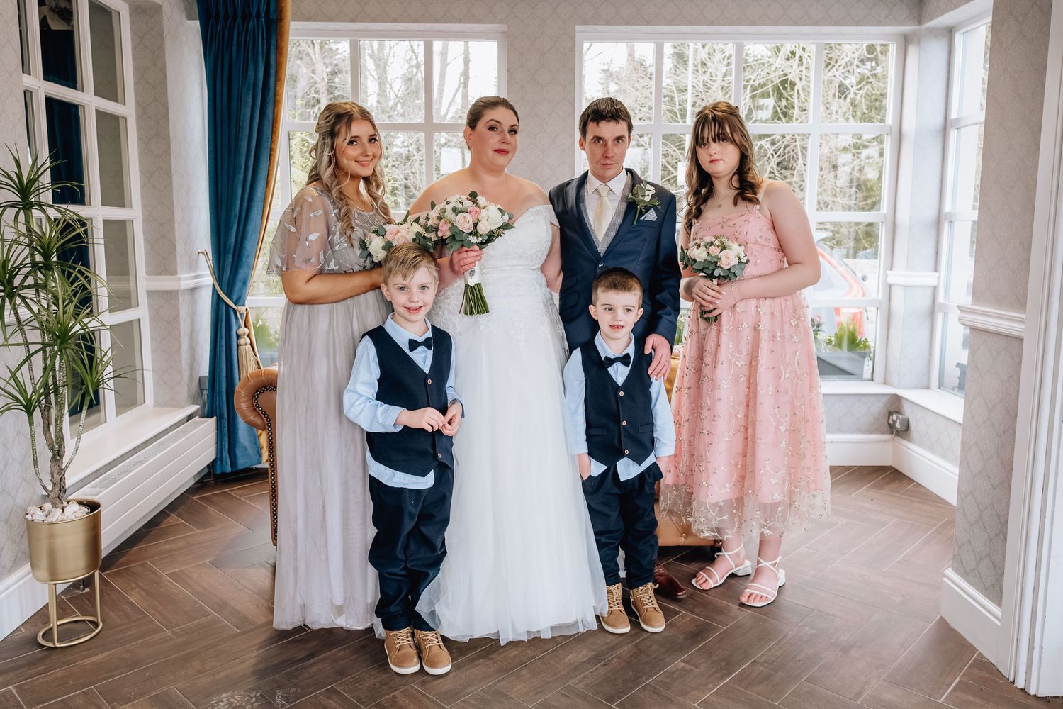 Wedding party portrait in sunlit room with bridesmaids in pink dresses and young ring bearers.
