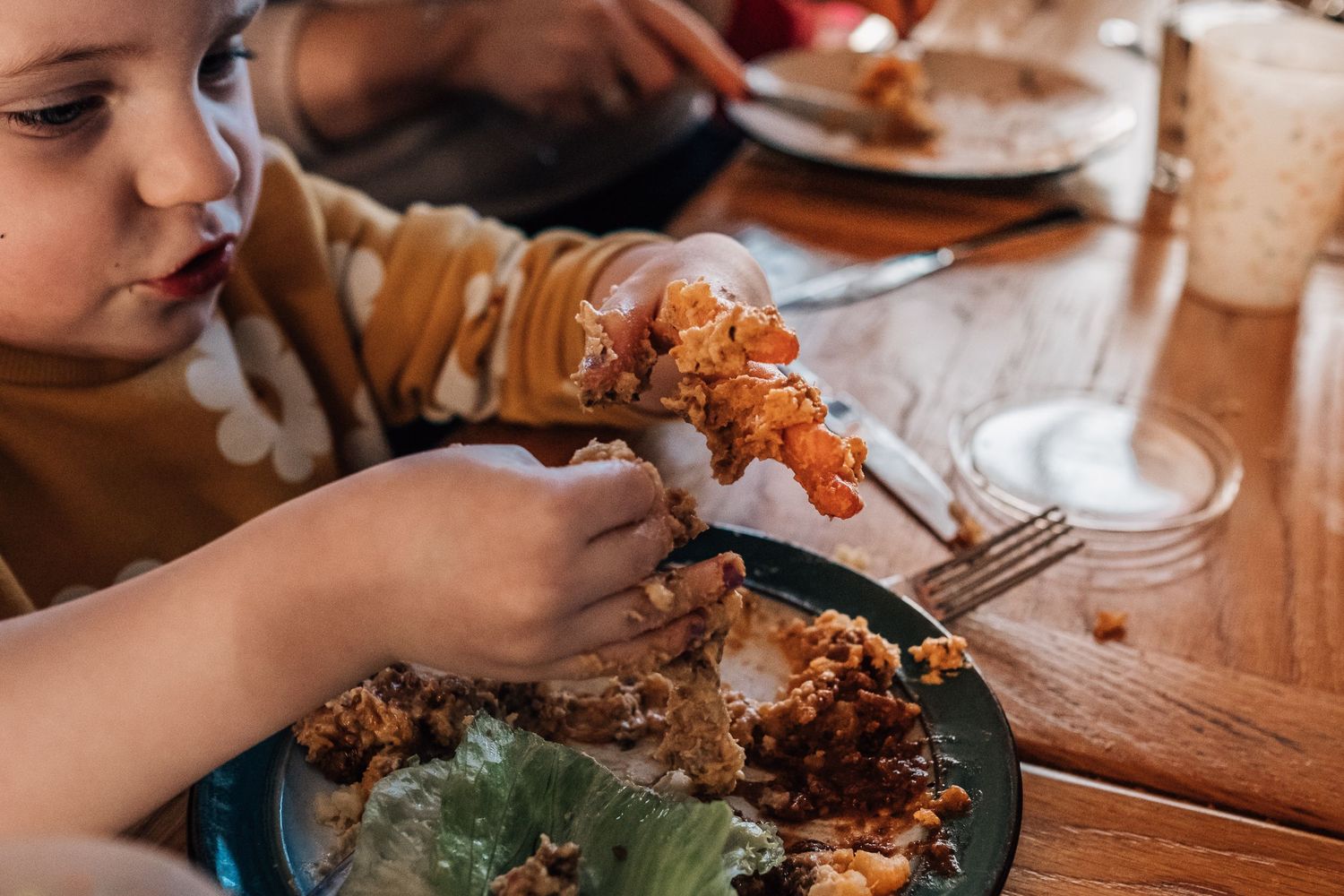 Small hands holding utensils while eating a meal with meat and vegetables on a dark plate.