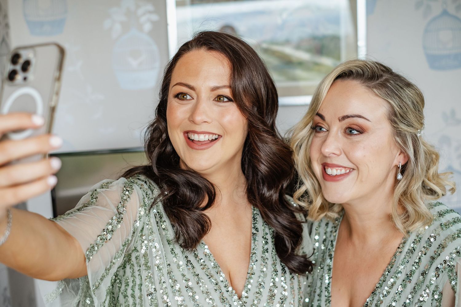 Two bridesmaids in mint green sequined dresses taking a selfie together during wedding preparations.