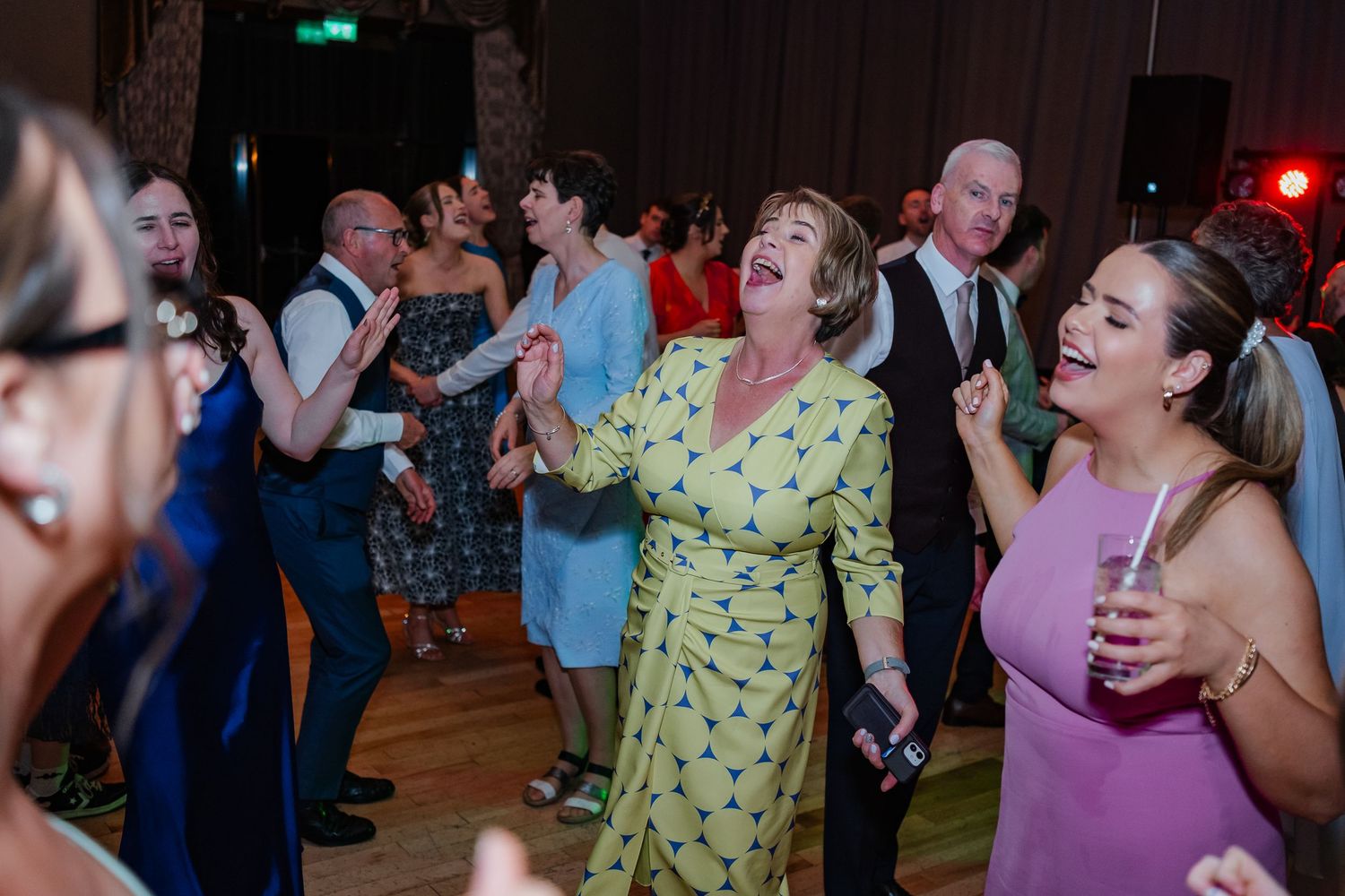 Wedding party guests dance and celebrate on a dimly lit dance floor with colored lighting.