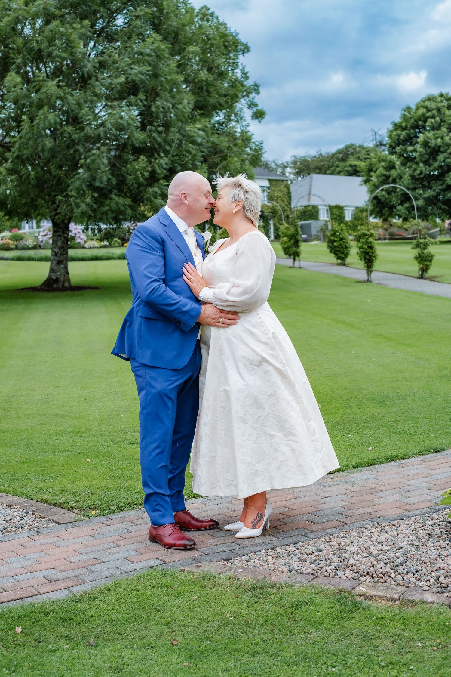 Bride and groom kissing in lush garden, bride in white tea-length dress, groom in royal blue suit.