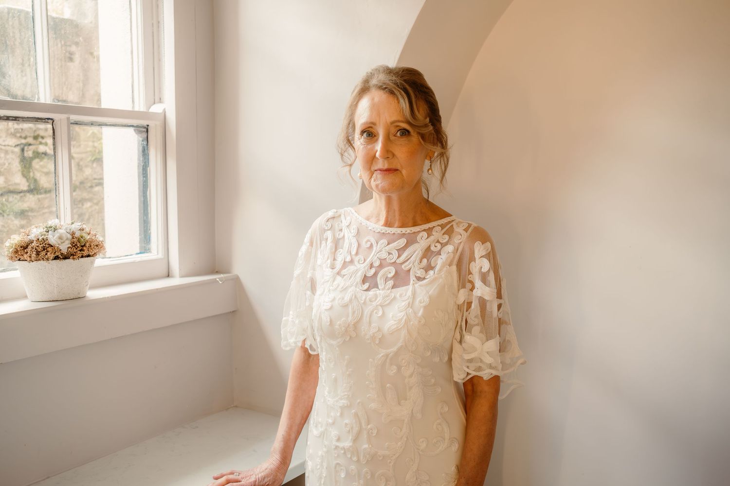 Bride in delicate lace dress poses by sunlit window in classic bridal portrait.