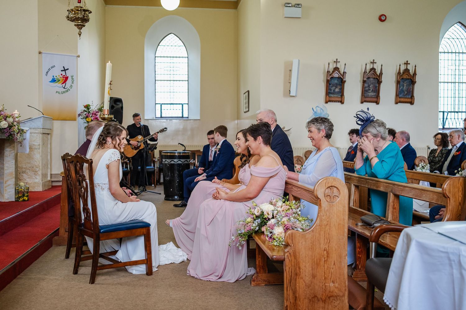 Wedding ceremony participants bow in prayer during traditional church service with wooden pews and stained glass windows.