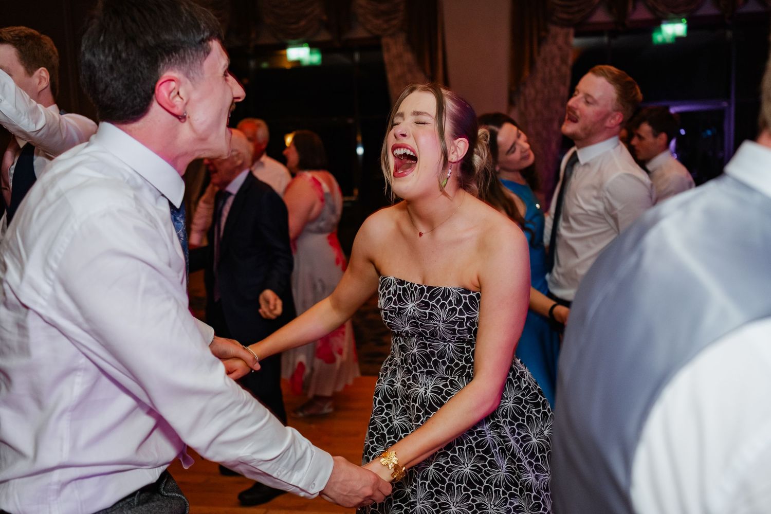 People dancing and celebrating at an energetic party or wedding reception with festive lighting in the background.