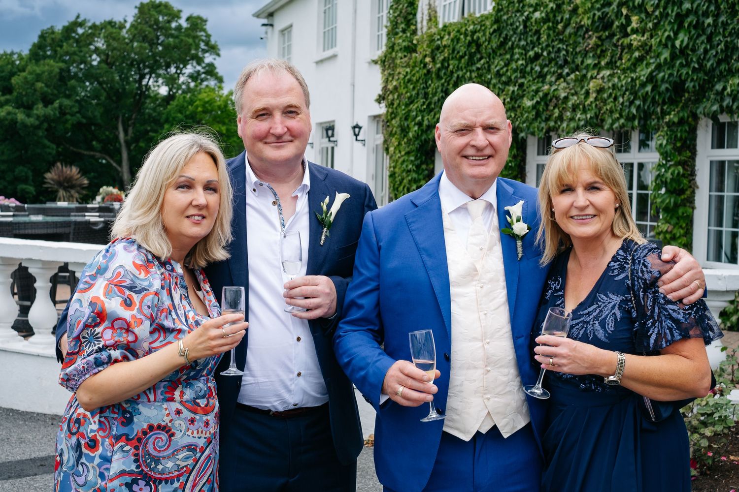 Two couples posing with champagne glasses outside elegant wedding venue with ivy walls.