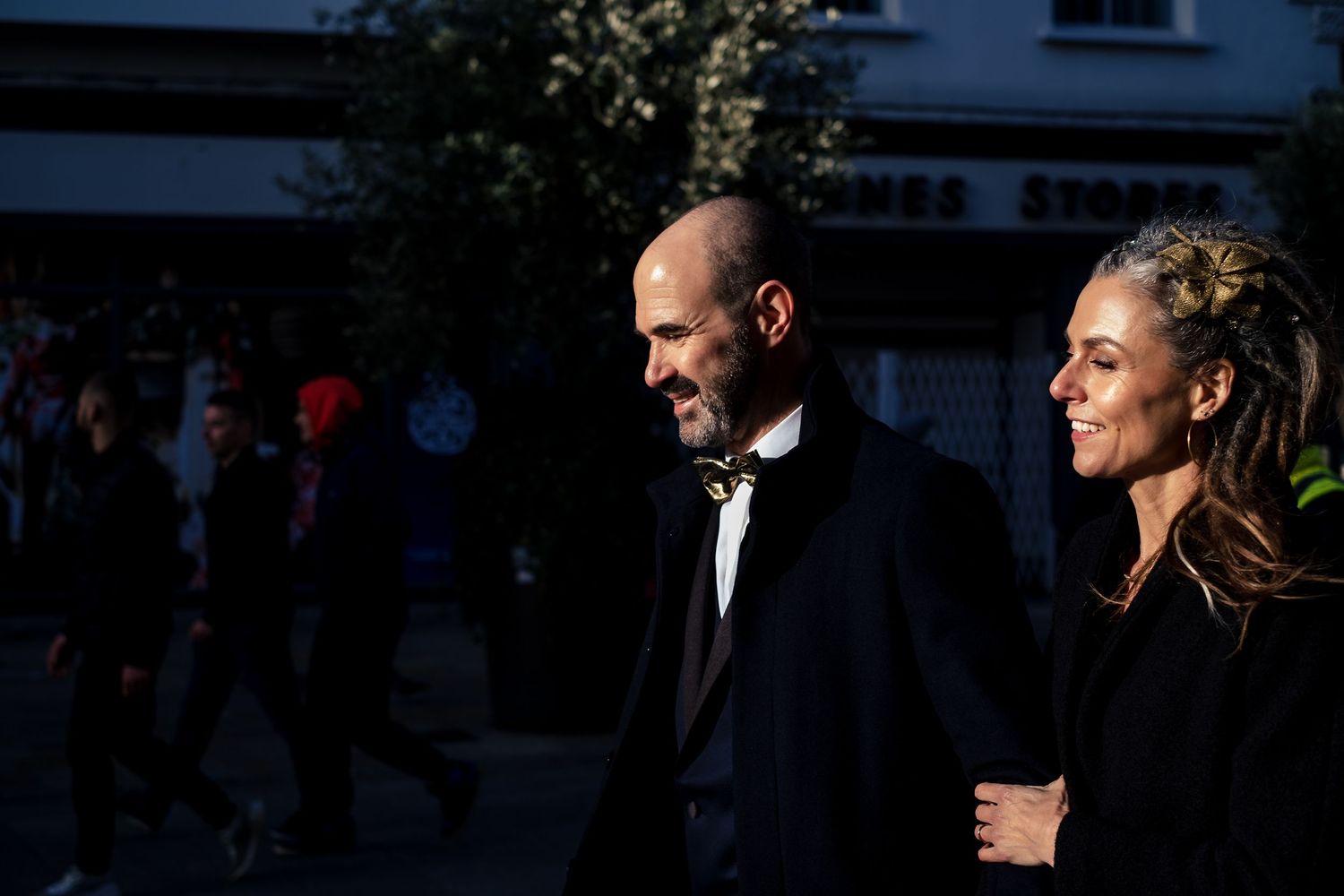 A bald man in a dark suit and a blonde woman walk together on a sunny street, smiling and enjoying each other's company.
