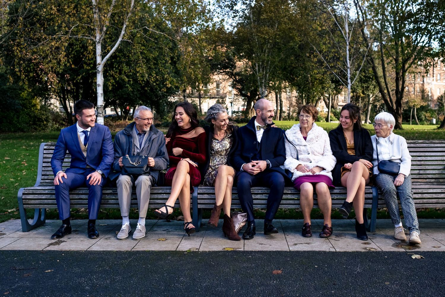 Seven adults sitting together on a park bench outdoors, smiling and socializing in a tree-lined urban garden.