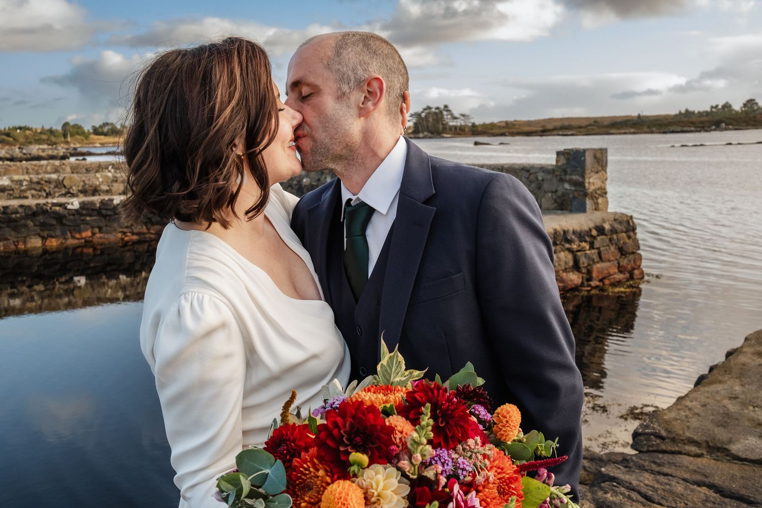 Bride and groom share a romantic kiss outdoors near a waterfront, bride holding a vibrant red and orange bouquet.
