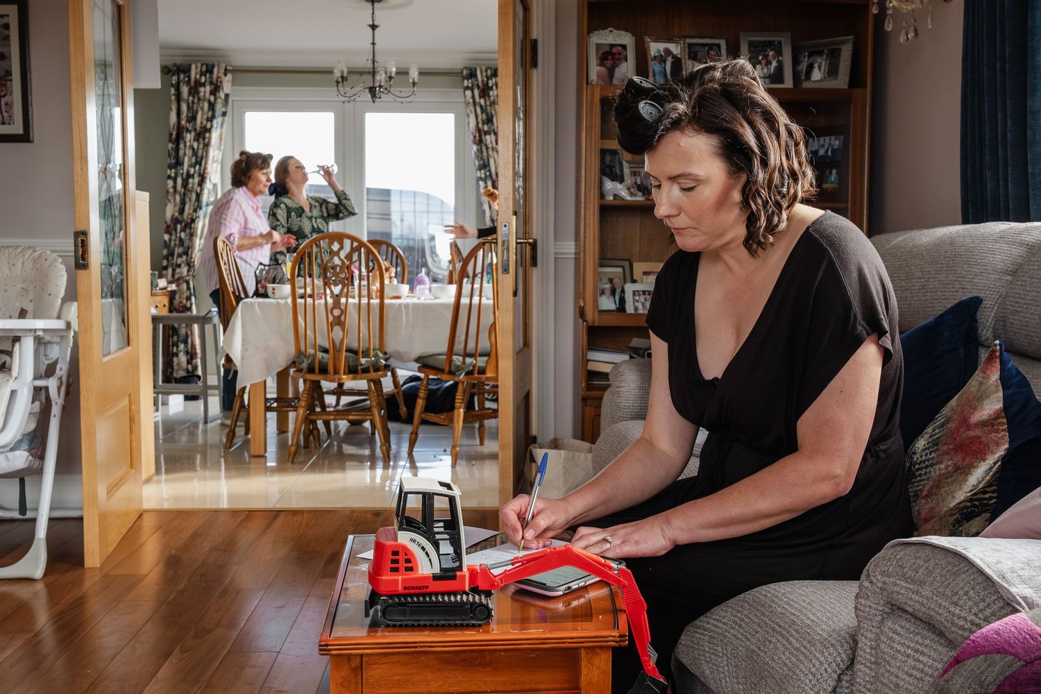 Woman sitting on sofa sewing red fabric while family gathers at dining table in background.