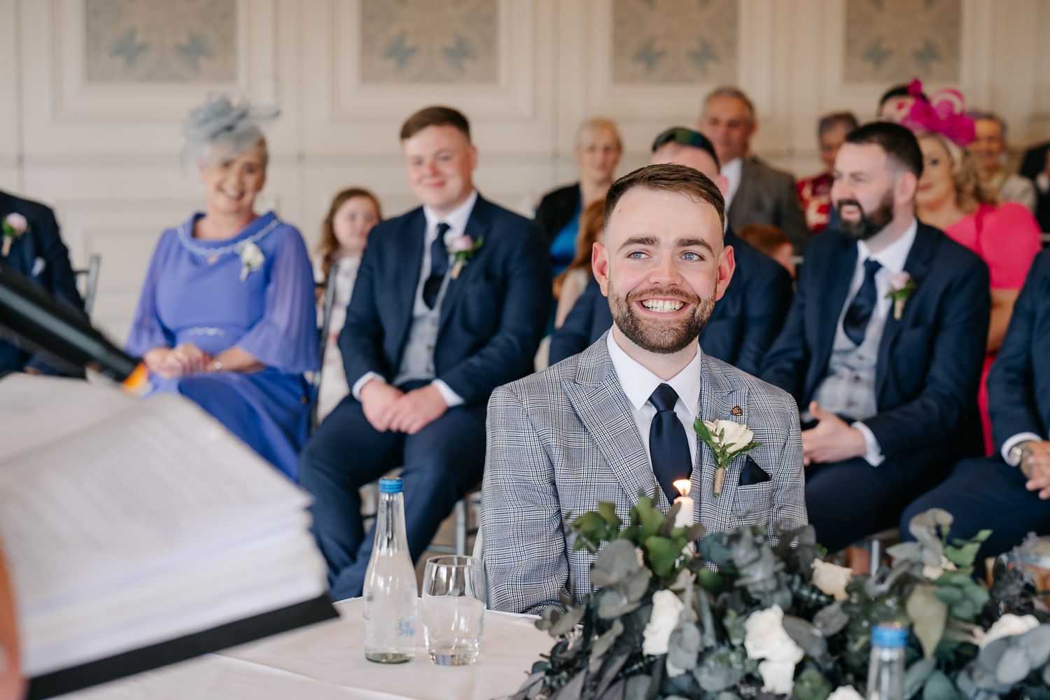 Smiling groom in grey suit sits at wedding ceremony table with guests seated behind him.