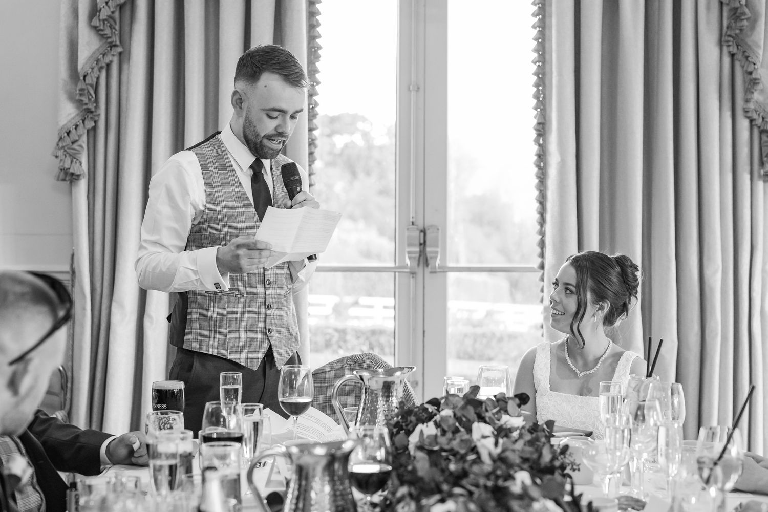 Groom reads wedding speech at elegant reception while bride smiles, black and white photography.