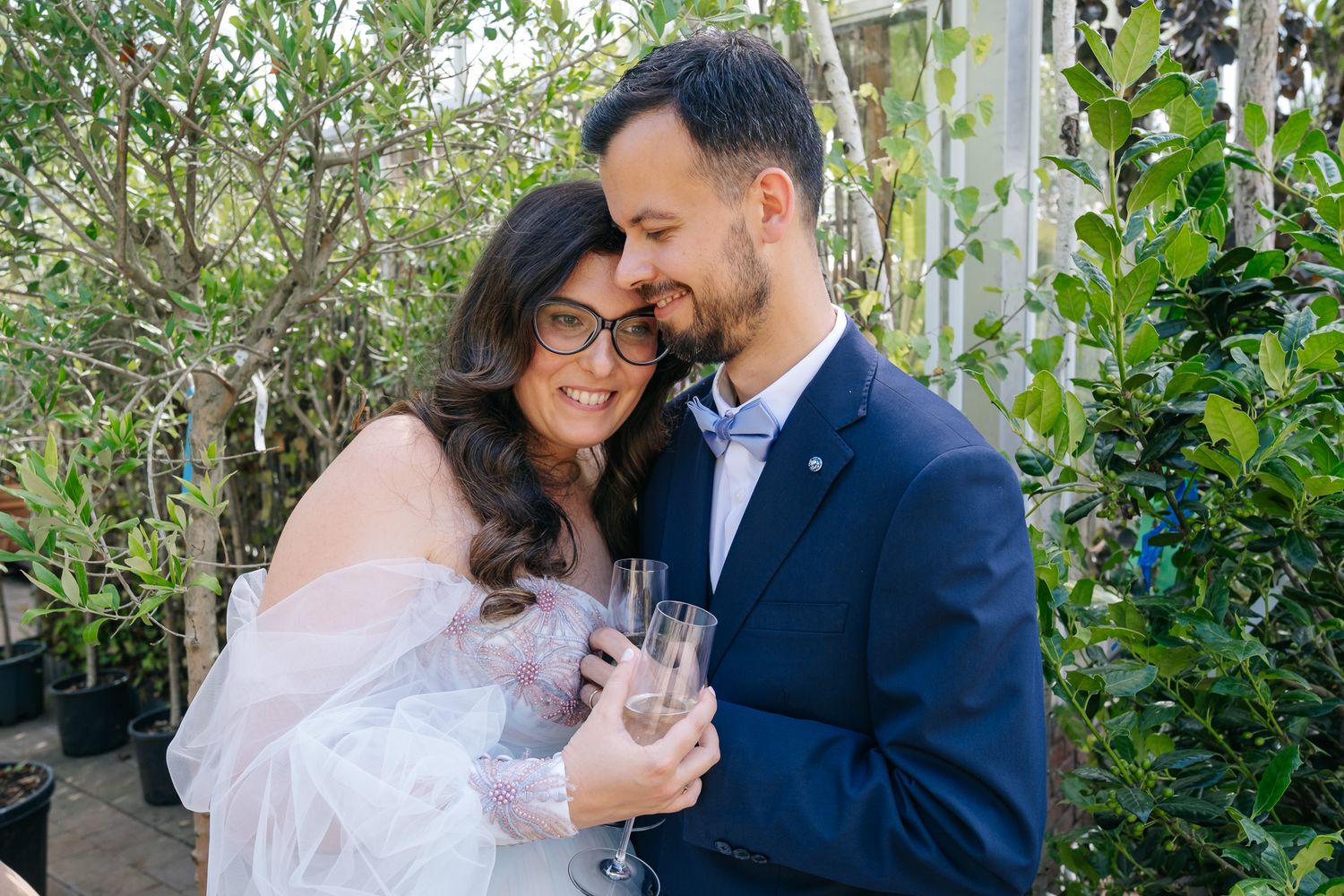 Couple in wedding attire embracing outdoors, bride in white veil and groom in navy suit holding champagne glasses.