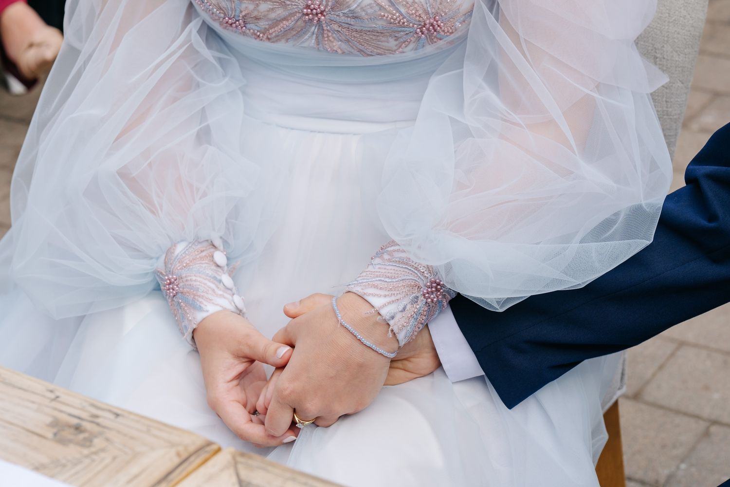 Bride and groom holding hands at wedding ceremony, bride wearing white tulle gown with lace sleeves, groom in navy suit.