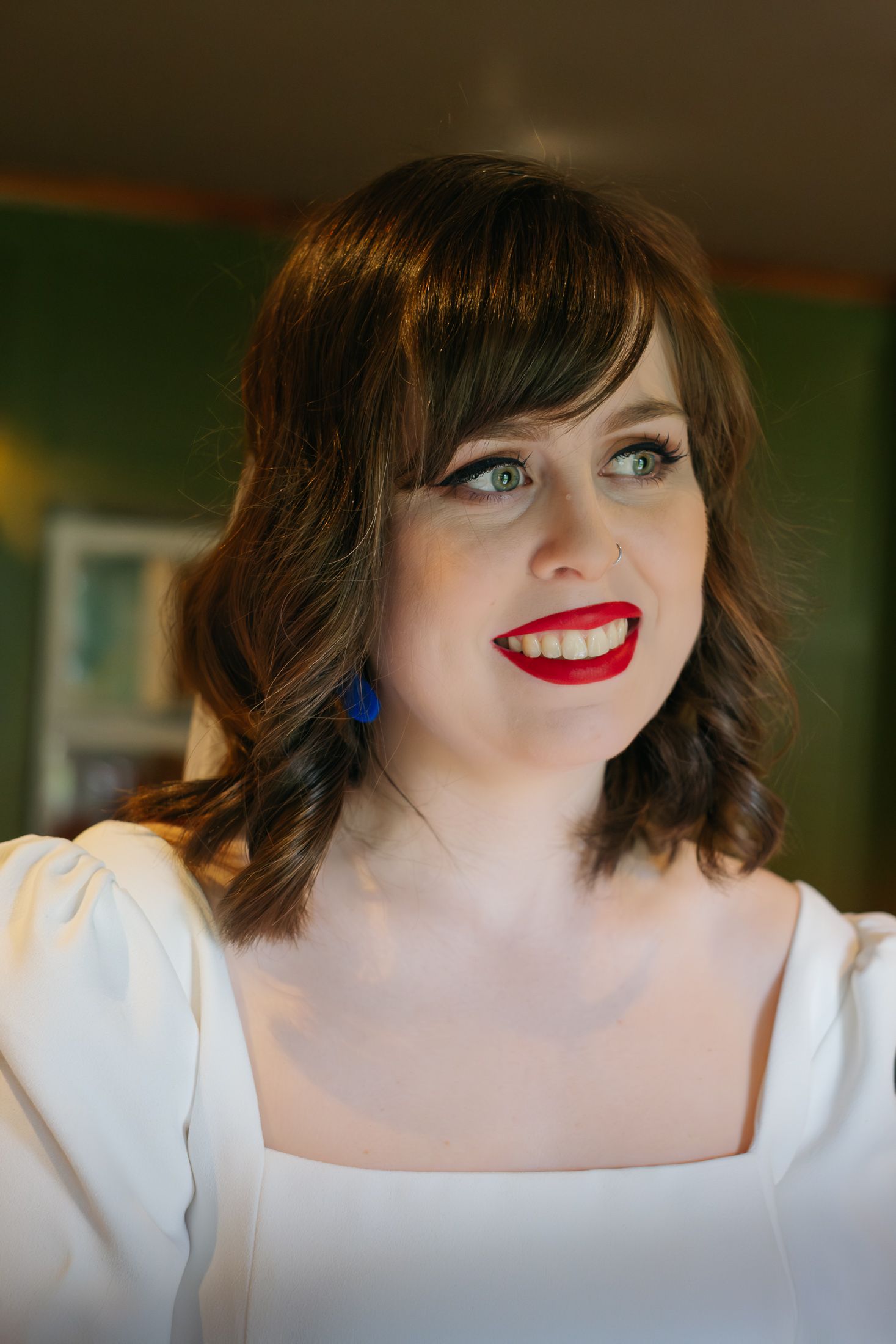 Smiling woman with red lips, blue earrings, and dark wavy hair wearing a white top in a warmly lit room.