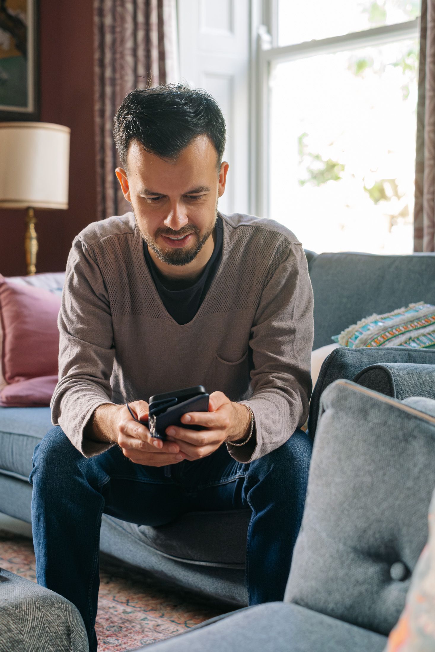 Man sitting on sofa using smartphone, smiling in a cozy living room with natural light from window.