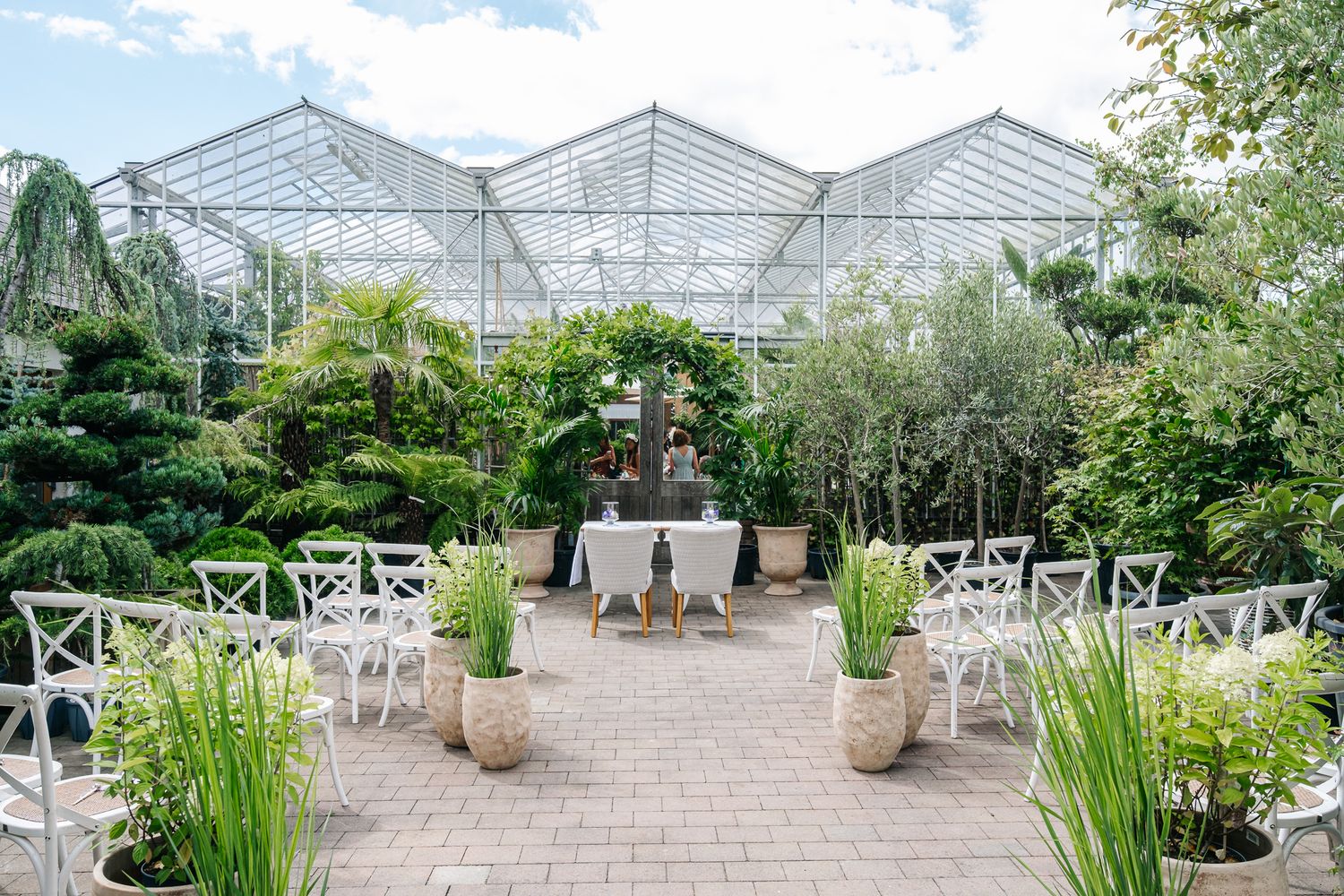Elegant greenhouse wedding venue with white chairs, lush greenery, and a draped table on a brick patio.