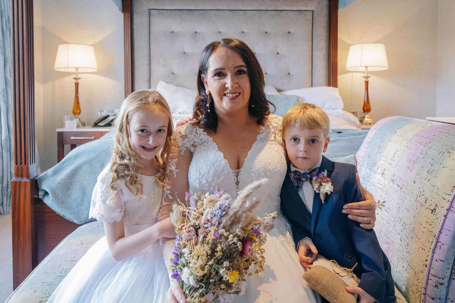 Bride in white wedding gown sits with flower girl and ring bearer in a decorated hotel bedroom.