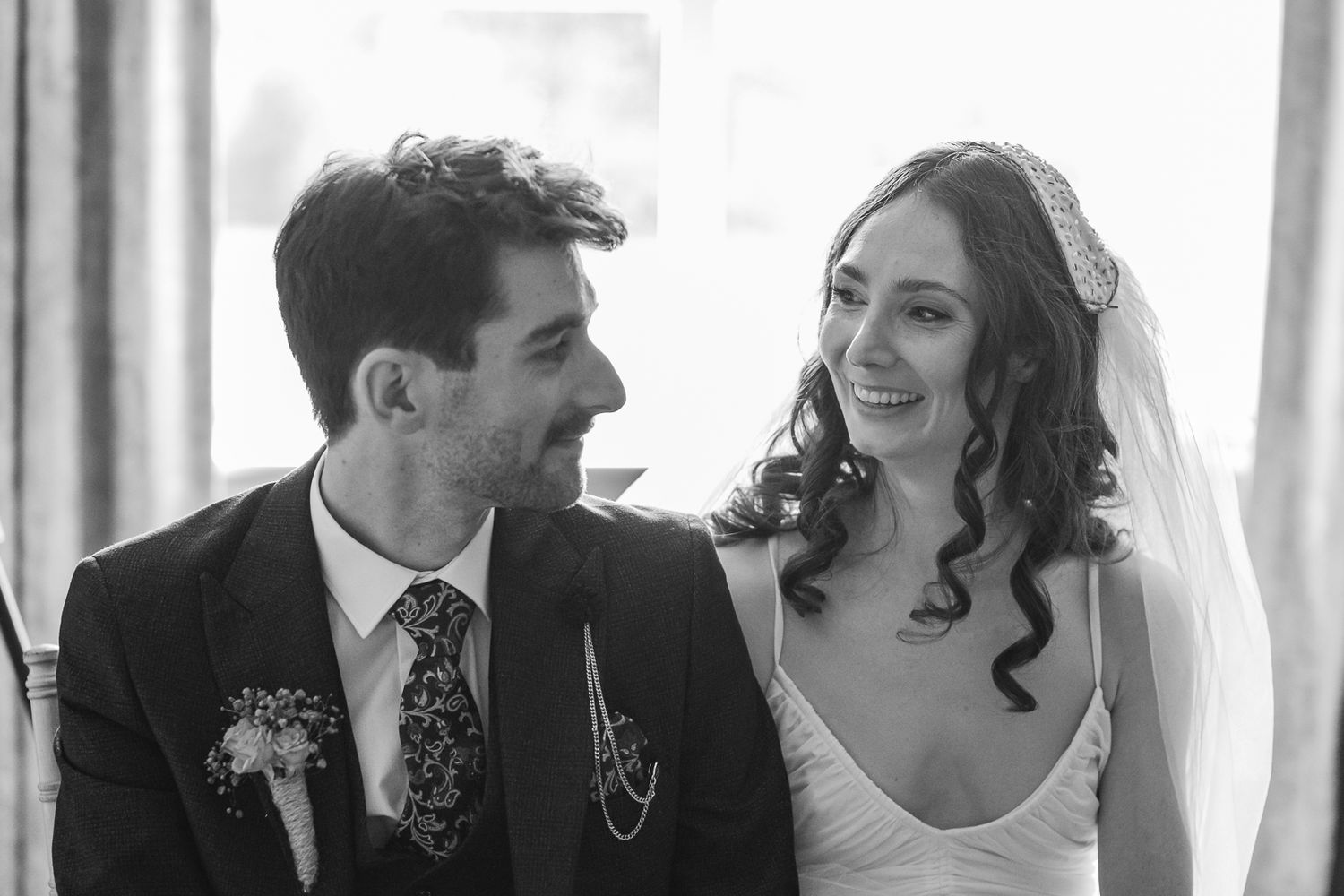 Black and white wedding photo of smiling bride in veil and groom in suit with floral tie gazing at each other.