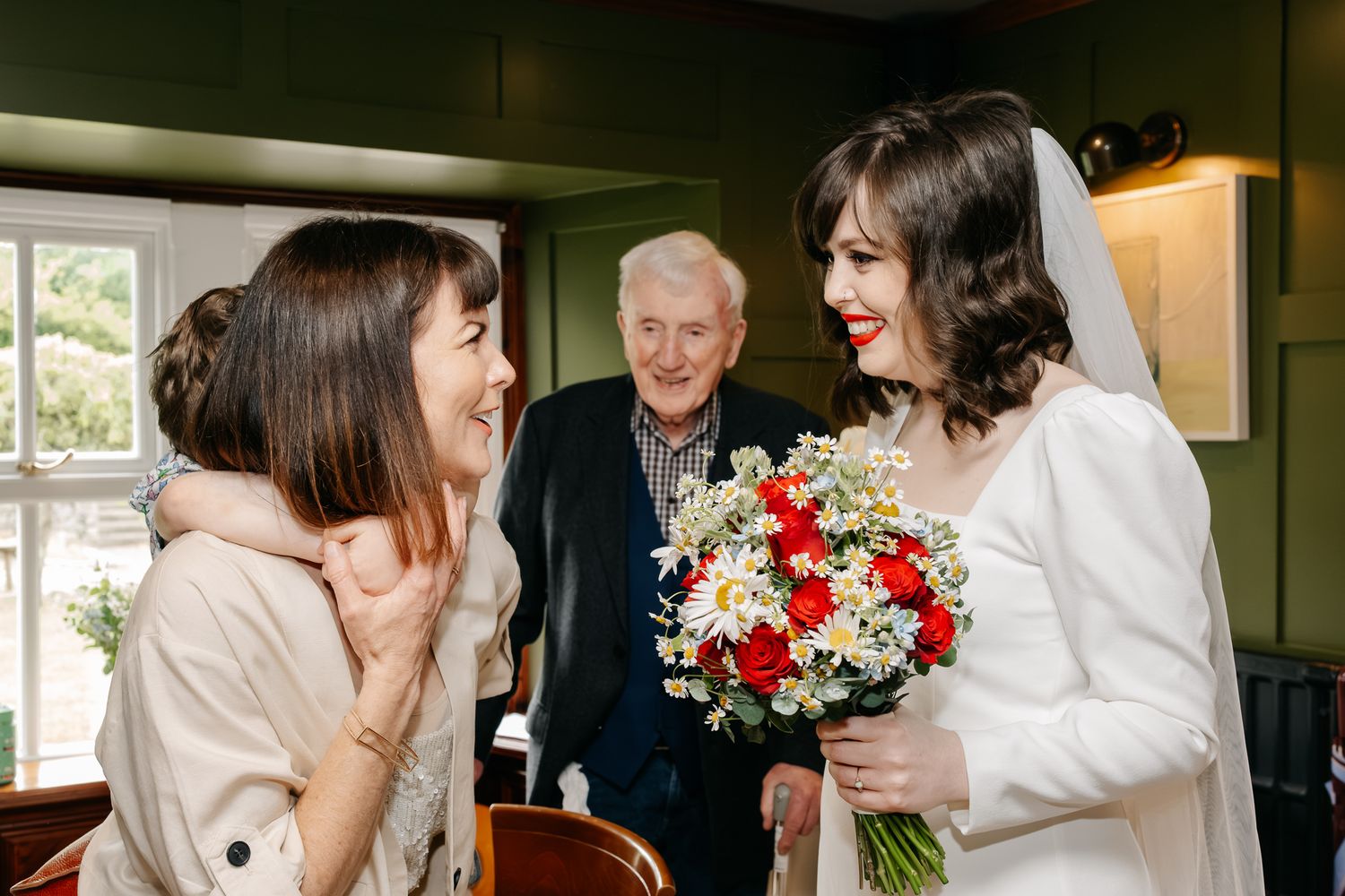 Bride in white gown holds colorful bouquet, smiling with guests at a wedding celebration indoors.