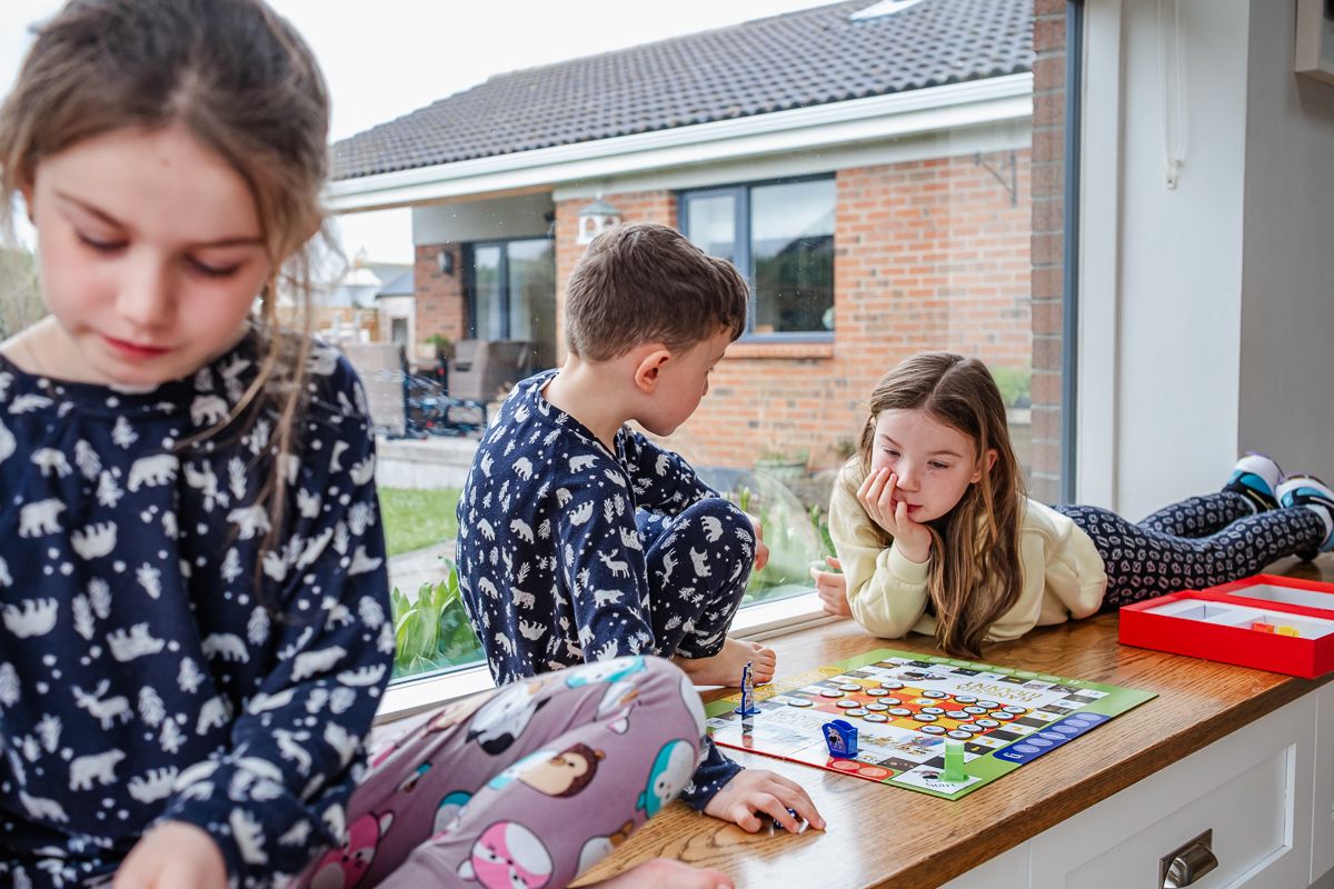 3 children quietly playing board games in a window seat in their home.
