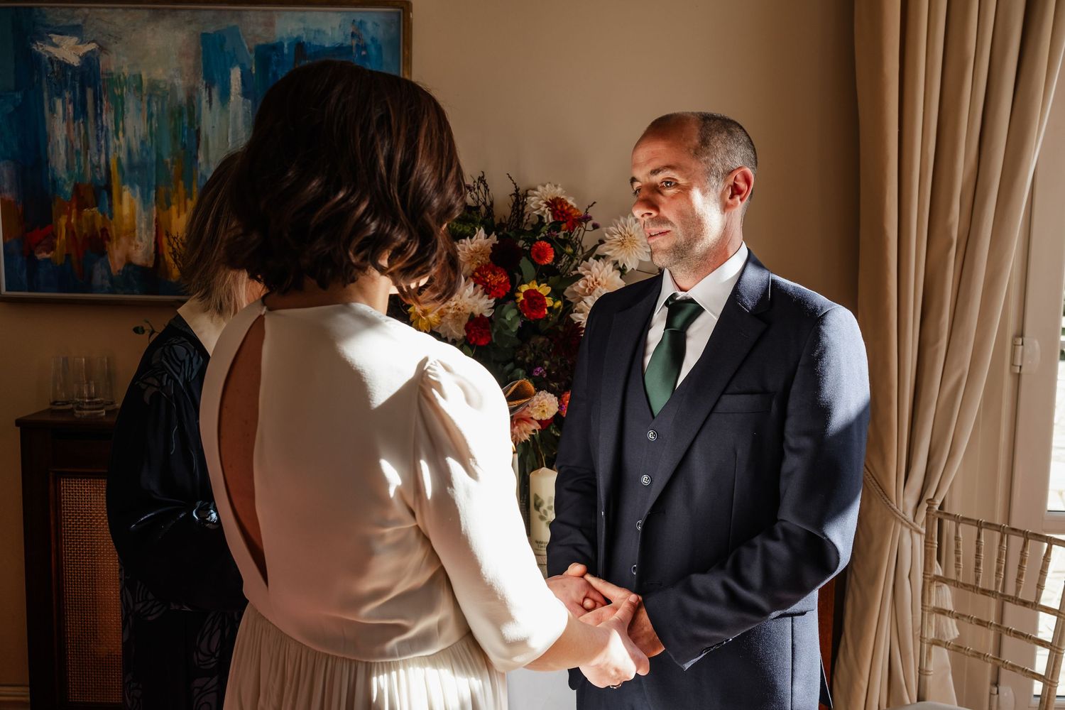 Groom in navy suit holds bride's hands during intimate wedding ceremony with floral arrangement in background.