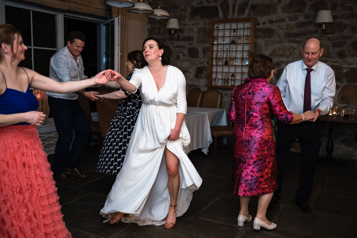 Bride in white gown dancing joyfully with guests at a rustic stone venue wedding reception.