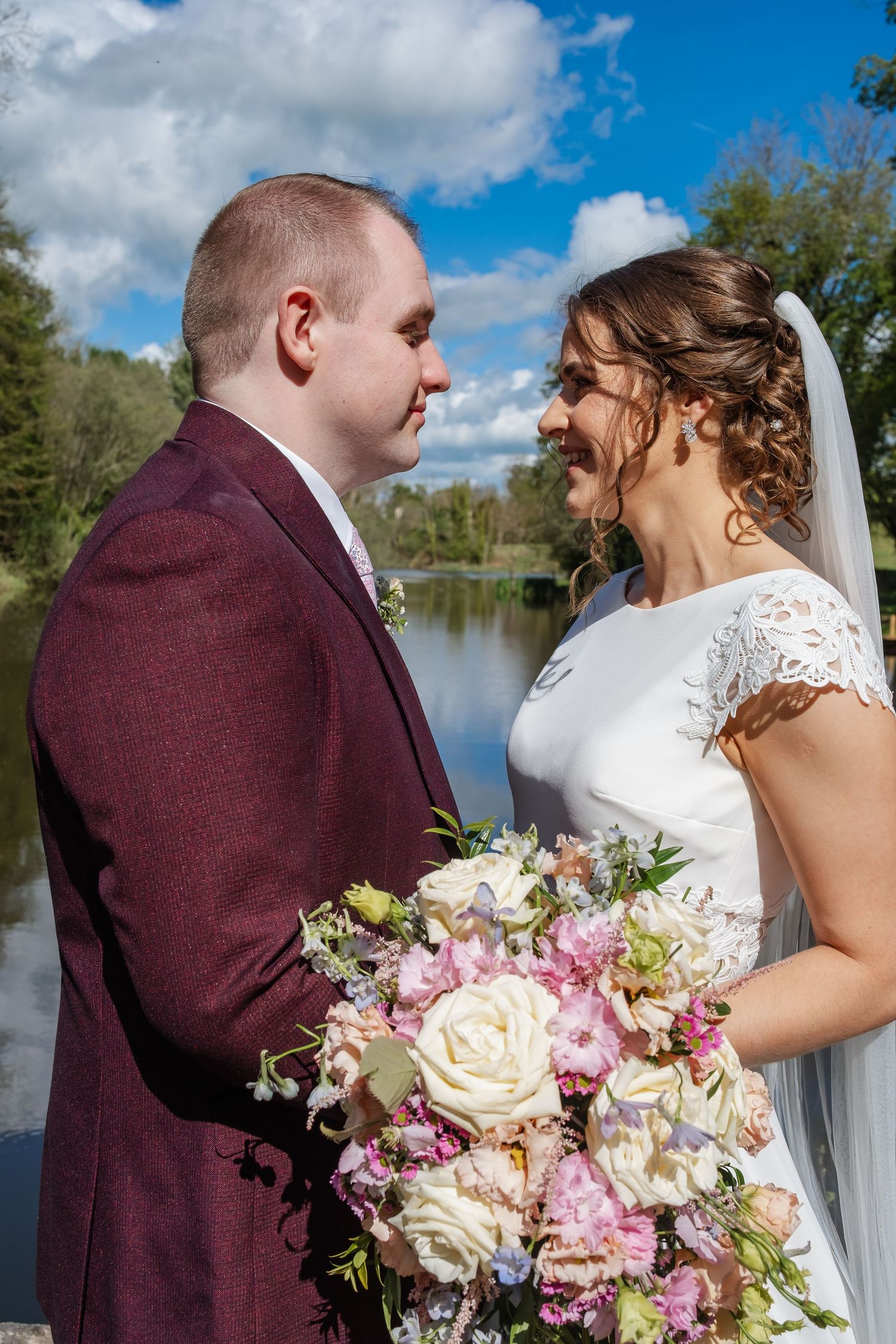 A couple shares a joyful moment by a lake on their wedding day with a stunning bouquet of pink and white roses.