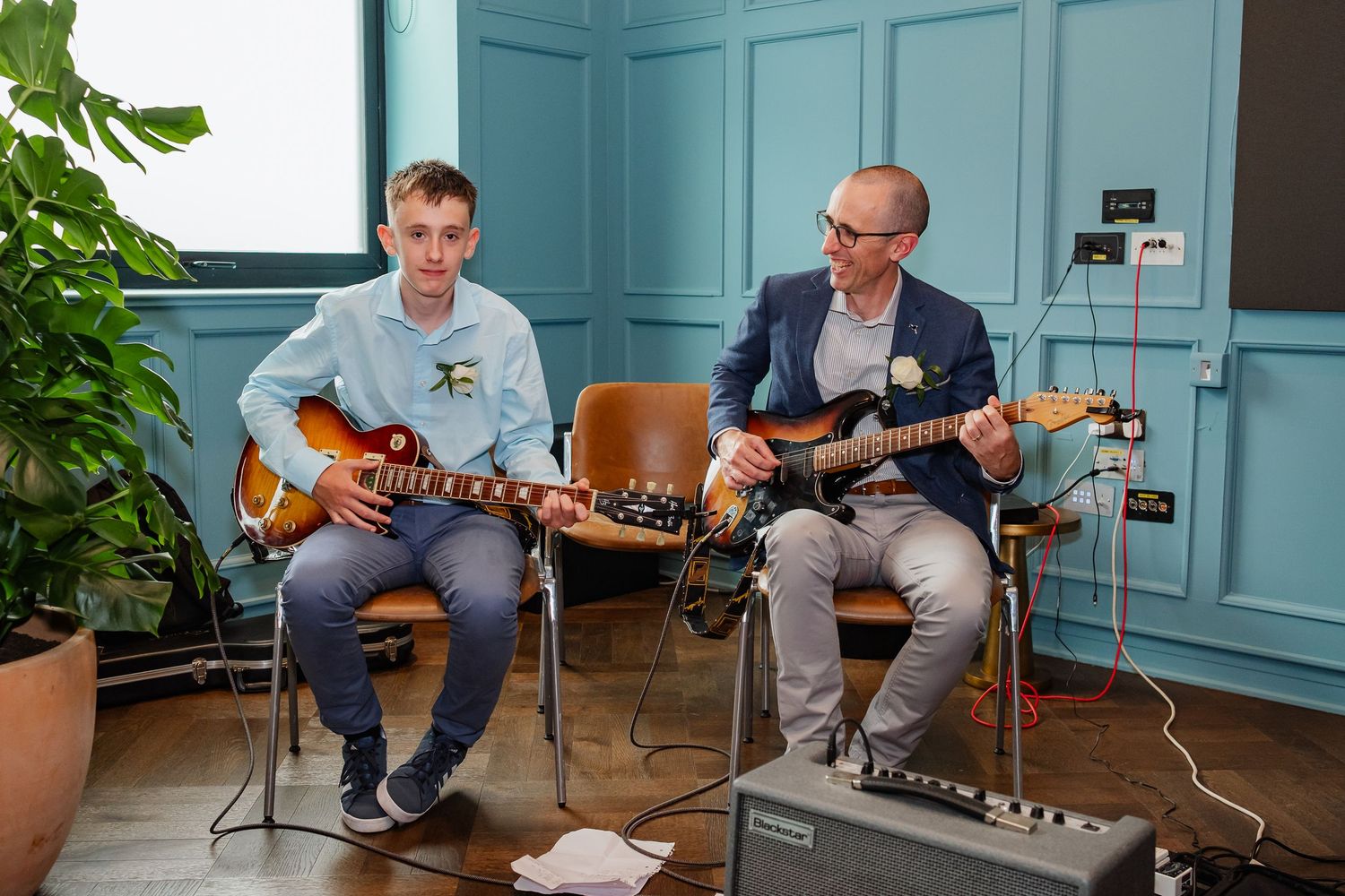 Musicians playing acoustic guitars in a room with teal blue walls and tropical plants.