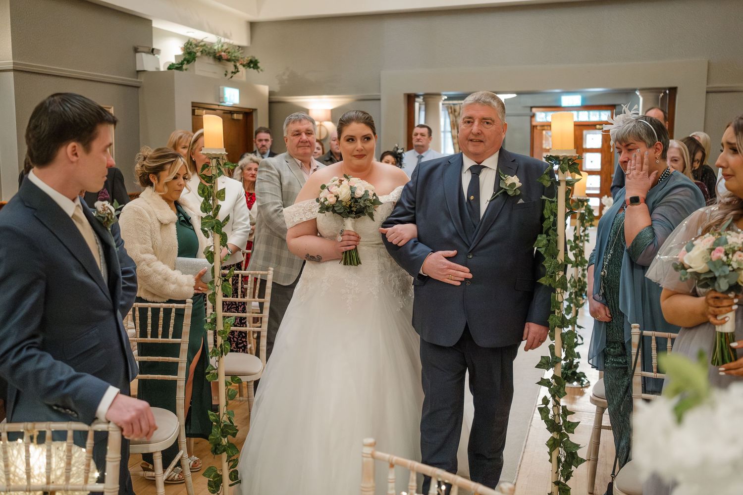 Wedding processional in an indoor venue decorated with floral arrangements and greenery.