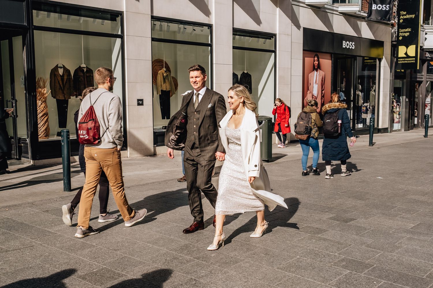 Elegantly dressed couple walking on a city street with storefronts and pedestrians.