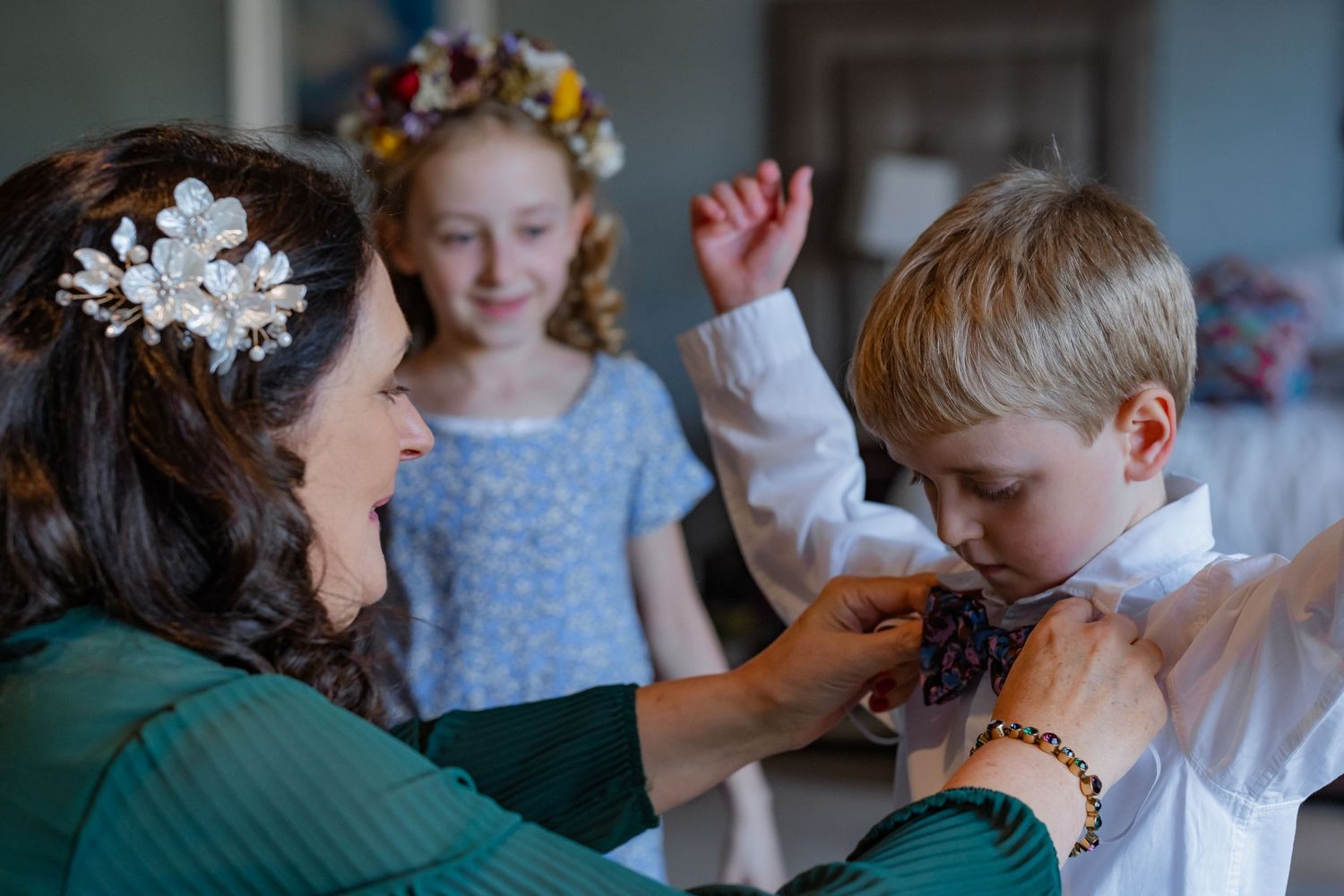 Woman adjusting boy's tie while girl in floral crown watches during wedding preparations.