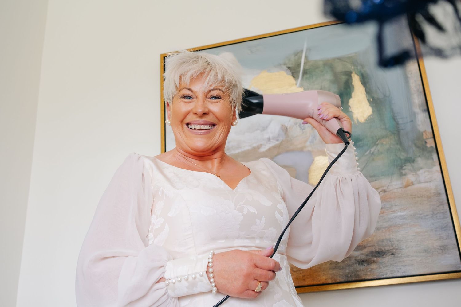 Smiling older woman in white wedding dress holding a microphone stand, posing in front of abstract gold artwork.