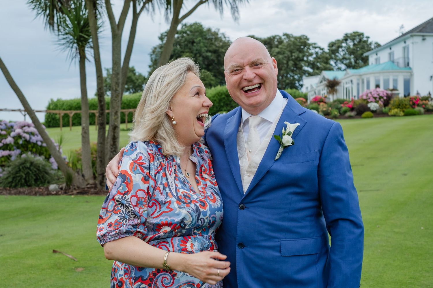 Joyful couple laughing outdoors at a wedding, man in blue suit with boutonniere, woman in floral dress.