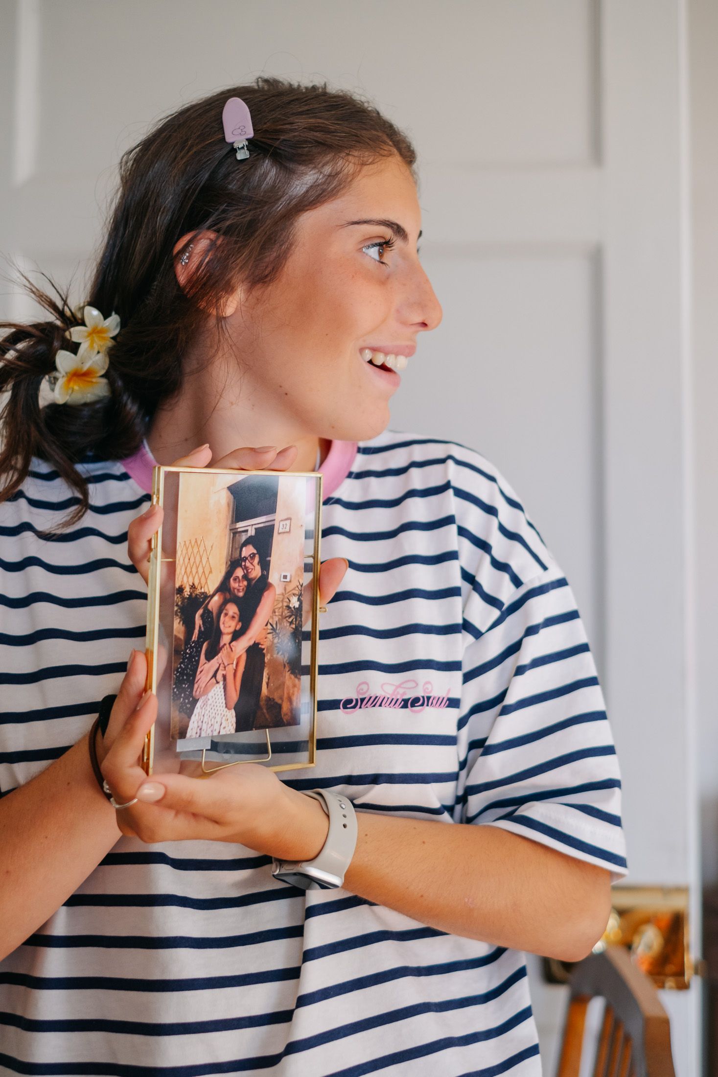 Smiling woman in striped shirt holding a framed photo, wearing a flower hair clip and smartwatch.