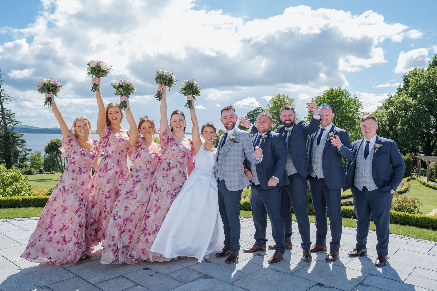 Wedding party posing outdoors, bridesmaids in pink floral dresses holding bouquets, groomsmen in navy suits, bride and groom centered.