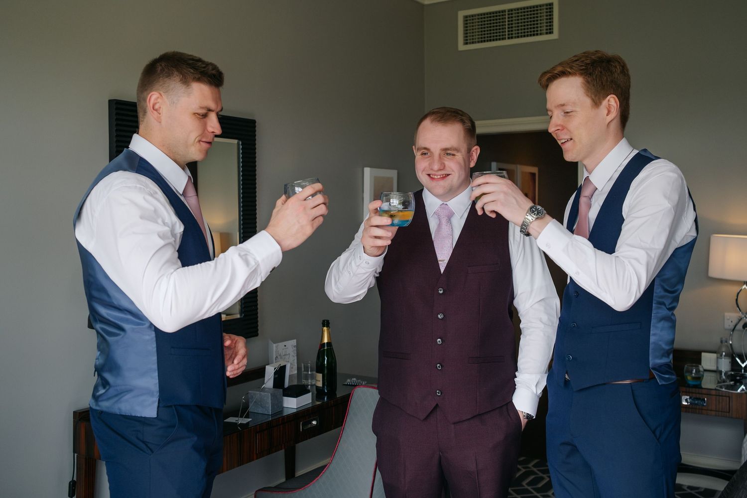 Groomsmen in navy and burgundy vests sharing drinks during wedding preparations.