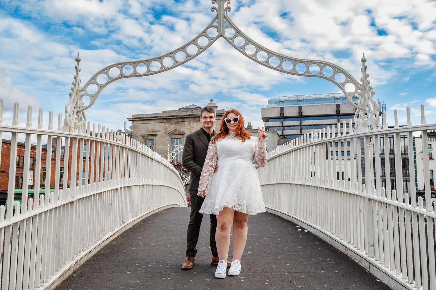Couple walking across Ha'penny Bridge in Dublin under cloudy blue sky.