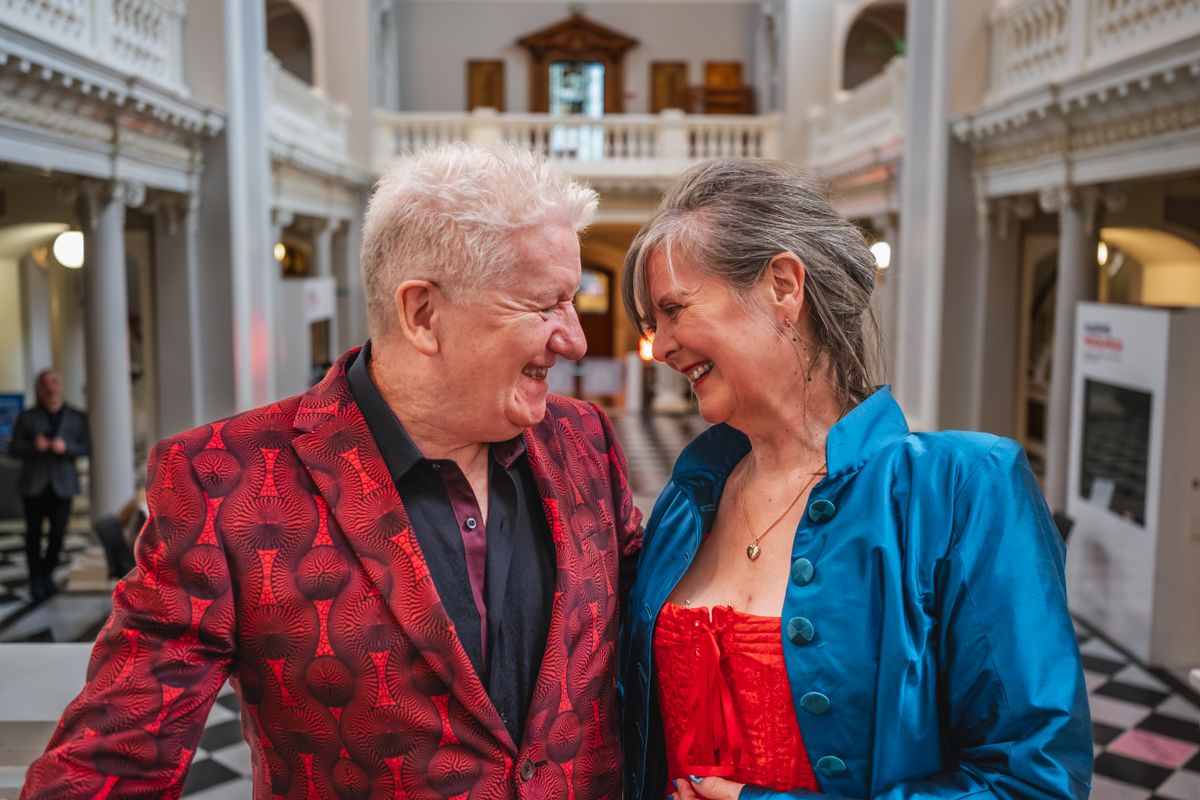 An elderly couple in matching red and blue outfits share a loving moment in an ornate indoor setting.