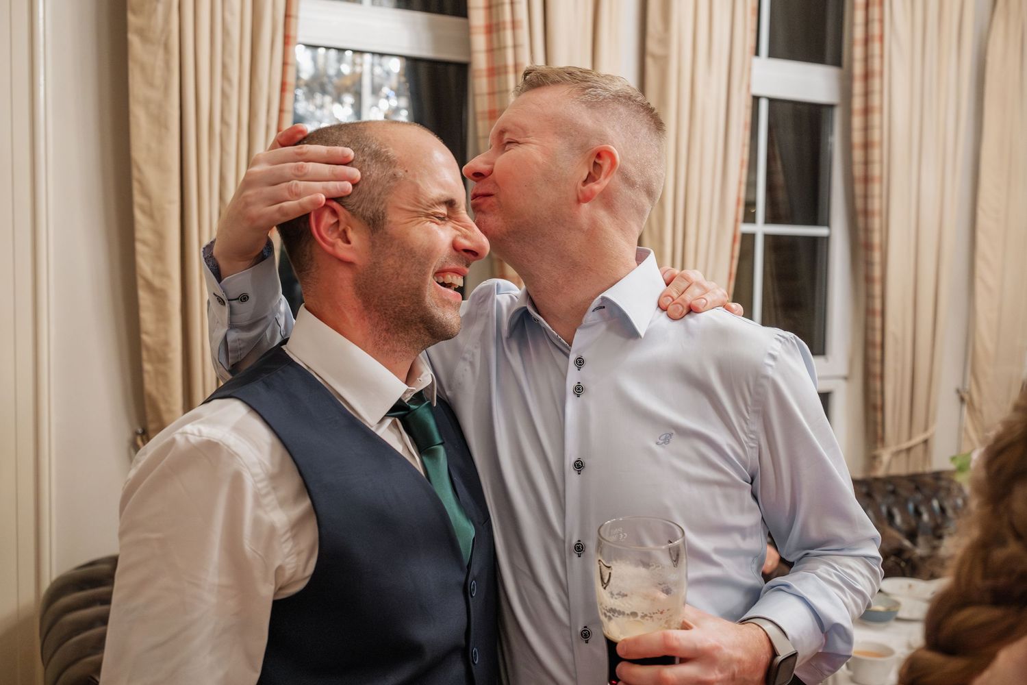 Two men share a joyful kiss and embrace at a wedding reception, one wearing a vest and green tie.